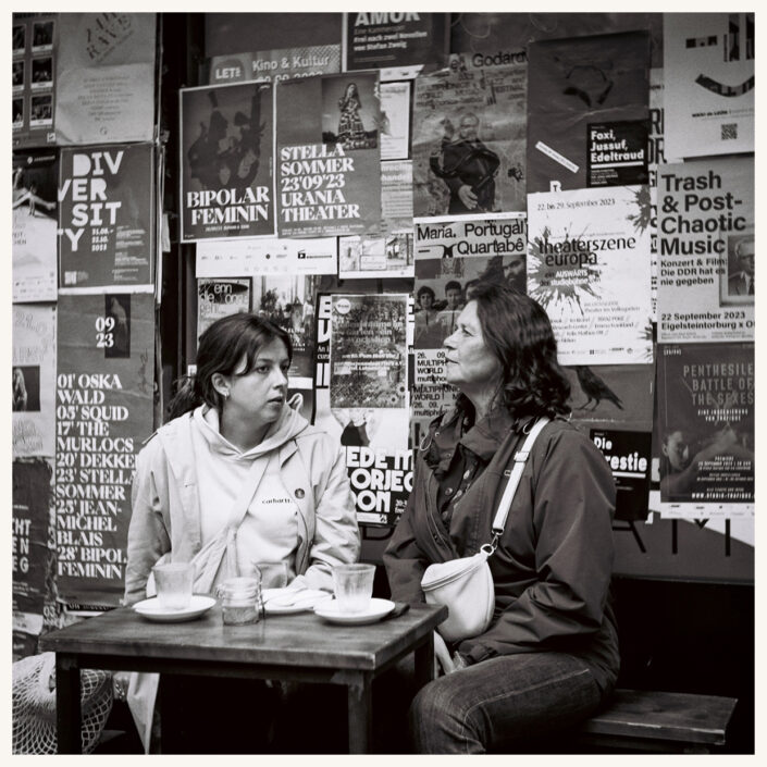 Zwei Frauen im Café vor einer mit Plakaten beklebten Wand, analog in Schwarz-Weiß fotografiert