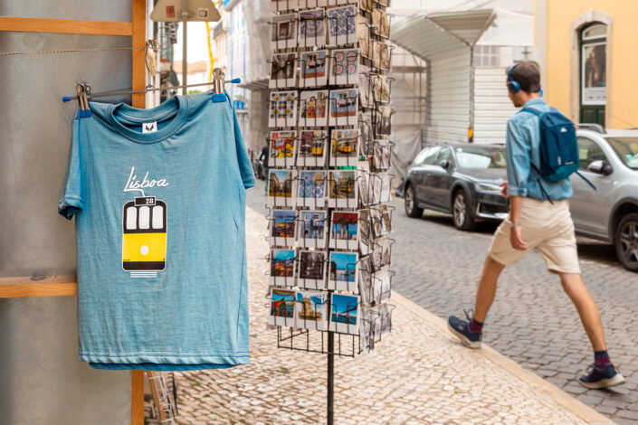 Ein Tourist geht an einem Souvenir Shop in Lissabon vorüber. Im Vordergrund ein Shirt mit dem Aufdruck Lisboa und der gelben Trambahn