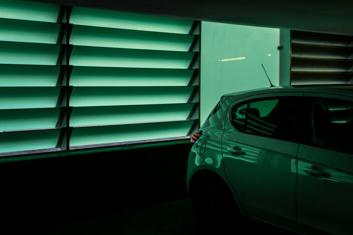 Ein geparktes Auto im Parkhaus, dahinter grüne grüne Lamellen, durch die Licht fällt