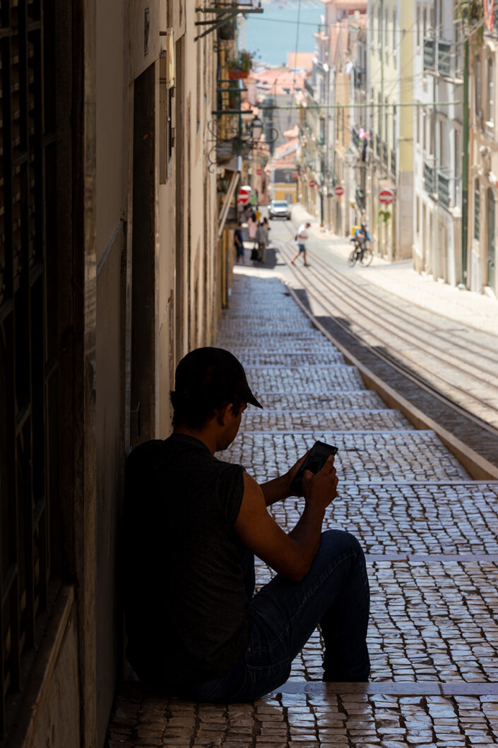 Ein junger Mann sitzt im Schatten auf einer Treppe, im Hintergrund eine typische Lissabonner Straße