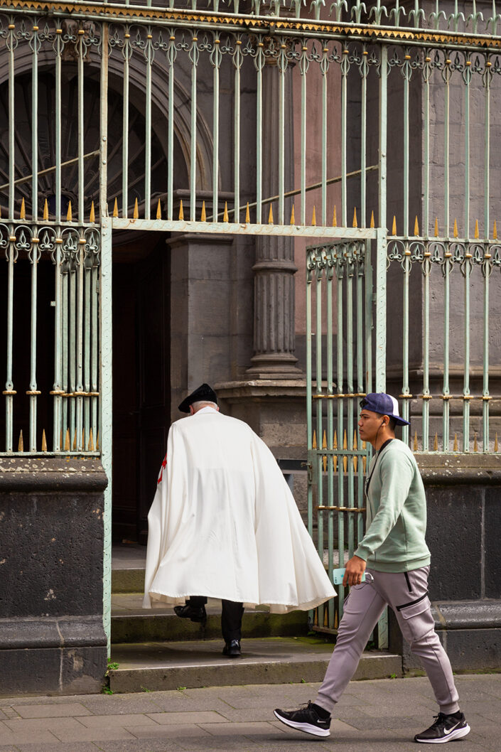 Ein Mann im weißen Umhang der durch das Tor zur Kirche Mariä Himmelfahrt und ein junger Mann, der vorüber geht