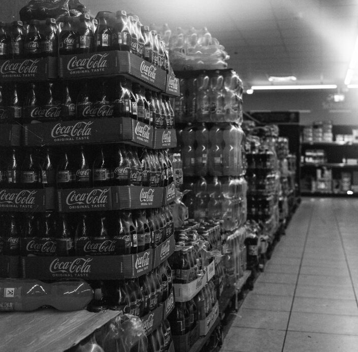 Ein Stapel Coca Cola Flaschen in einem Supermarktgang, analog in Schwarz-Weiß fotografiert